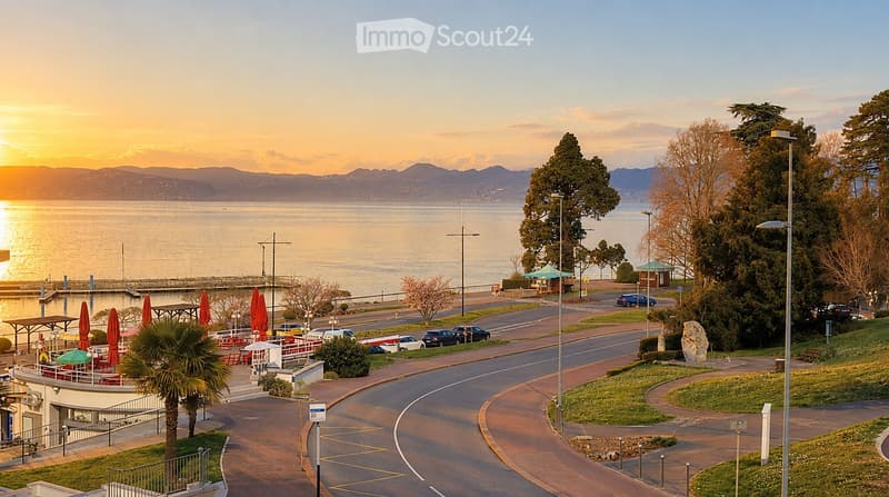 Coastal promenade with a view of the ocean at sunset, featuring palm trees, closed umbrellas, parked cars, and a winding road.