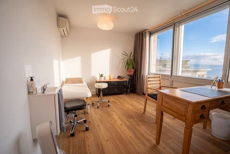 Bright, modern room with wooden flooring, large windows offering a sea view, a desk with a chair, a small table with a stool, a plant, and a mirror on the wall.