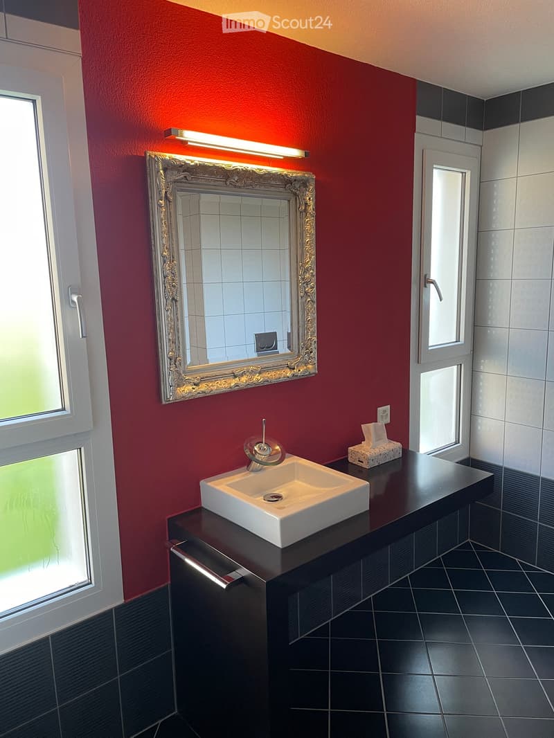 Modern bathroom with red accent wall, white sink, chrome faucet, ornate mirror, and black tiled floor.