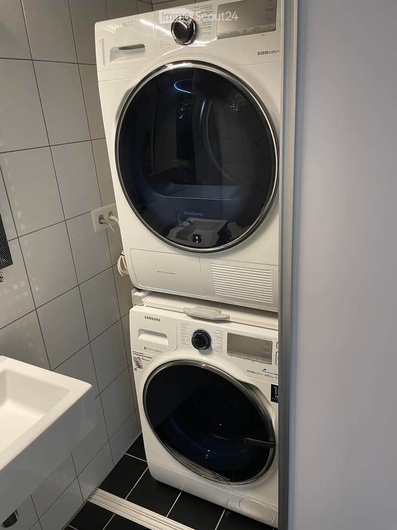 Modern bathroom with a white Samsung washing machine and a white Samsung dishwasher stacked vertically, grey tiled walls, and a white sink.