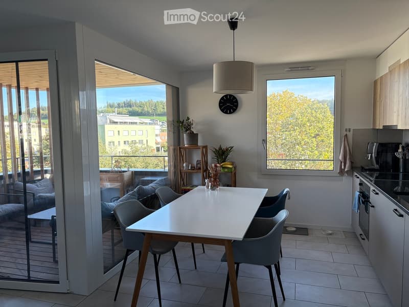 Modern open-plan living and dining area with a white dining table, grey chairs, a grey sofa, a clock on the wall, and a kitchen area with cabinets and appliances. Large windows and a sliding glass door lead to a balcony with outdoor seating.