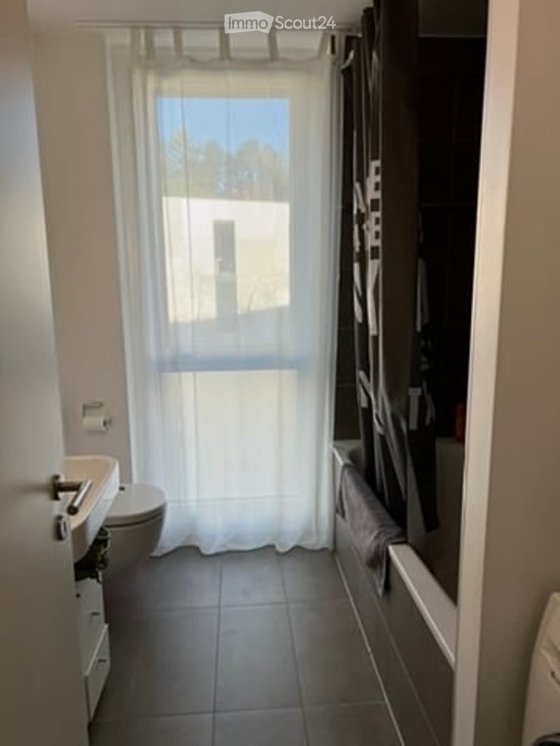 Modern bathroom with a white sink, toilet, bathtub, and a large window with a white curtain. The floor is tiled, and the walls are painted white.