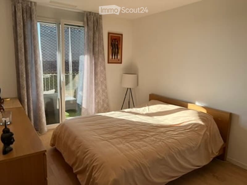 A modern bedroom with a large bed, nightstand, floor lamp, and a picture frame on the wall. The room has large windows with blinds and curtains, leading to a balcony.