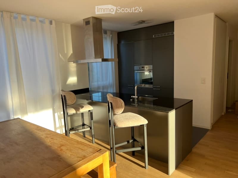 Modern kitchen with dark cabinets, stainless steel appliances, a central island with bar stools, and a wooden dining table in the foreground.