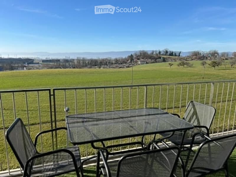 Balcony with a metal railing, overlooking a green field and distant hills. A glass-top table and four metal chairs are placed on the balcony.