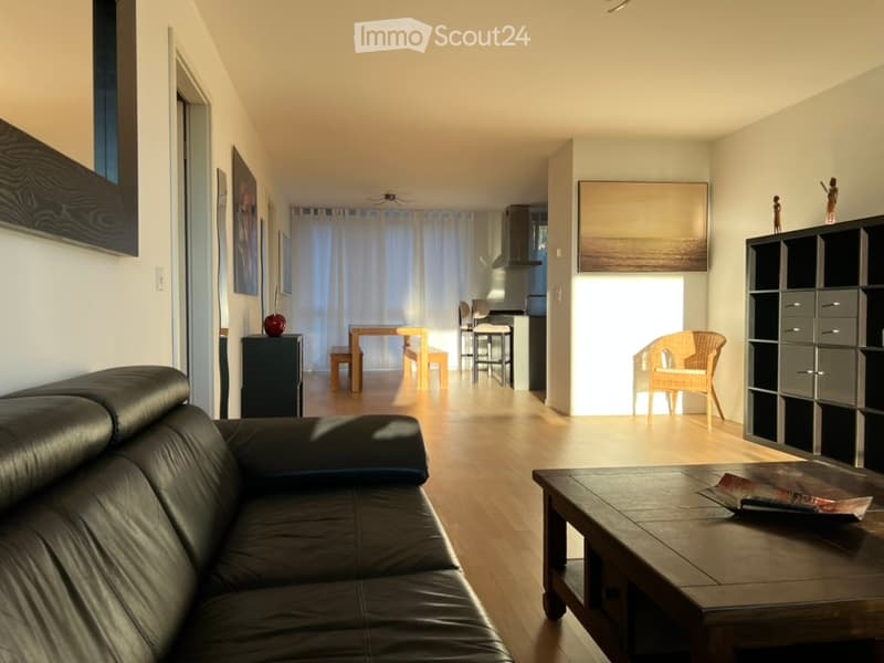 A modern, open-concept living and dining area with a black leather sofa, wooden coffee table, dining table with chairs, and a small kitchen area visible in the background.