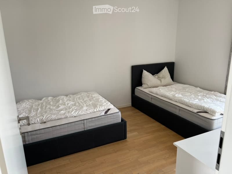 A small bedroom with two single beds, each with white bedding and pillows, placed on a wooden floor against white walls.