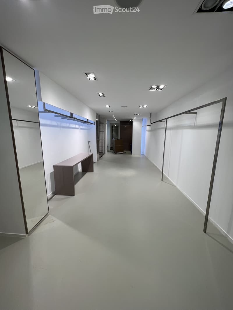Modern, minimalist hallway with white walls, grey carpeted floor, and recessed lighting. Features a long bench on the left side and a large mirror on the wall.