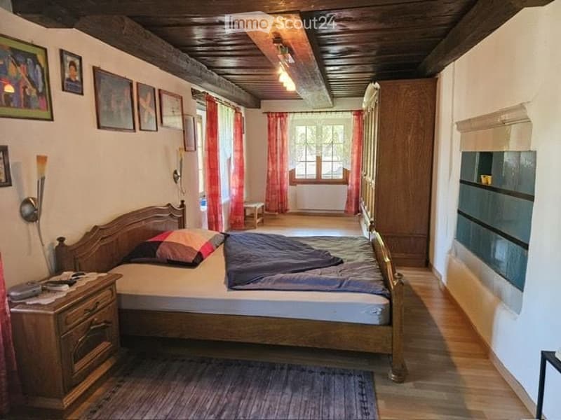 Cozy bedroom with wooden bed, nightstand, lamp, picture frames, and a small table near the window with red curtains.