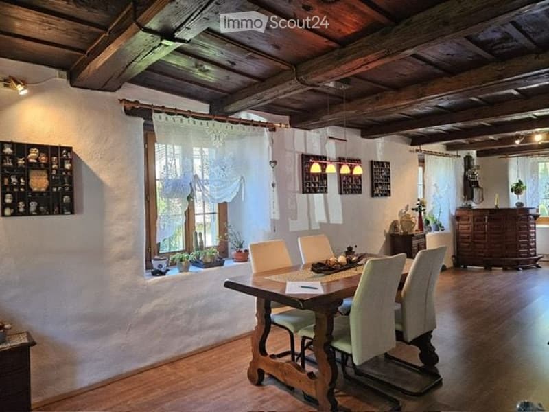 Cozy dining room with exposed wooden beams, a large wooden dining table surrounded by six chairs, wall-mounted shelves, and potted plants on the windowsill.