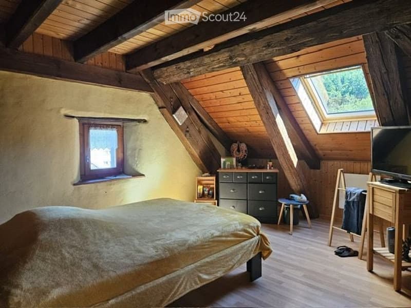 Cozy attic bedroom with wooden beams, slanted ceilings, and exposed wooden beams. Features a large bed, nightstand, chest of drawers, small stool, mirror, television, and desk.