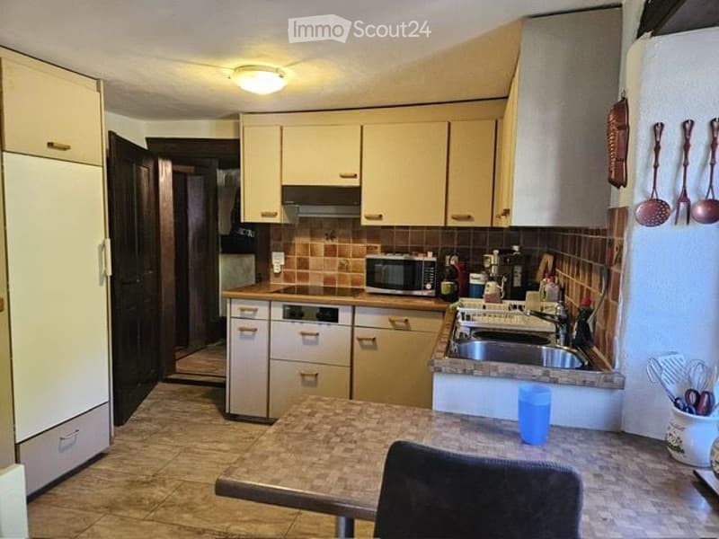 A well-equipped kitchen with light-colored cabinets, a stainless steel sink, a microwave, and a refrigerator. The countertop features a blue cup and a chair is positioned near the counter.