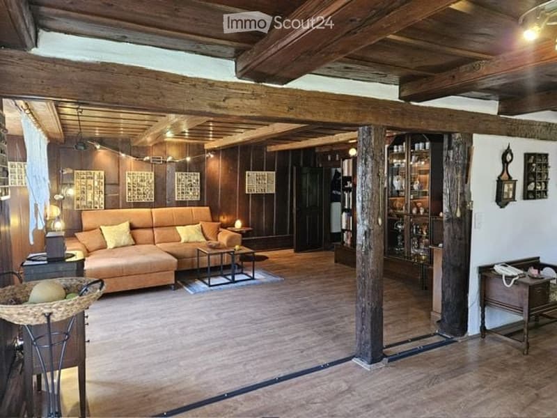 Cozy living room with exposed wooden beams, wooden flooring, and warm lighting. Features a large orange sectional sofa with yellow pillows, a coffee table, side tables, a bookshelf, and a telephone on a small table.