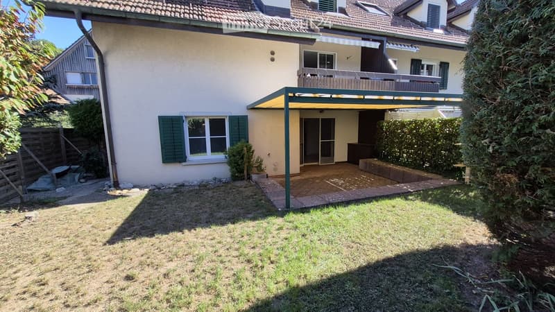 Backyard with a covered patio area, lawn, and surrounding greenery. The house has beige walls, green shutters, and a balcony on the upper floor.