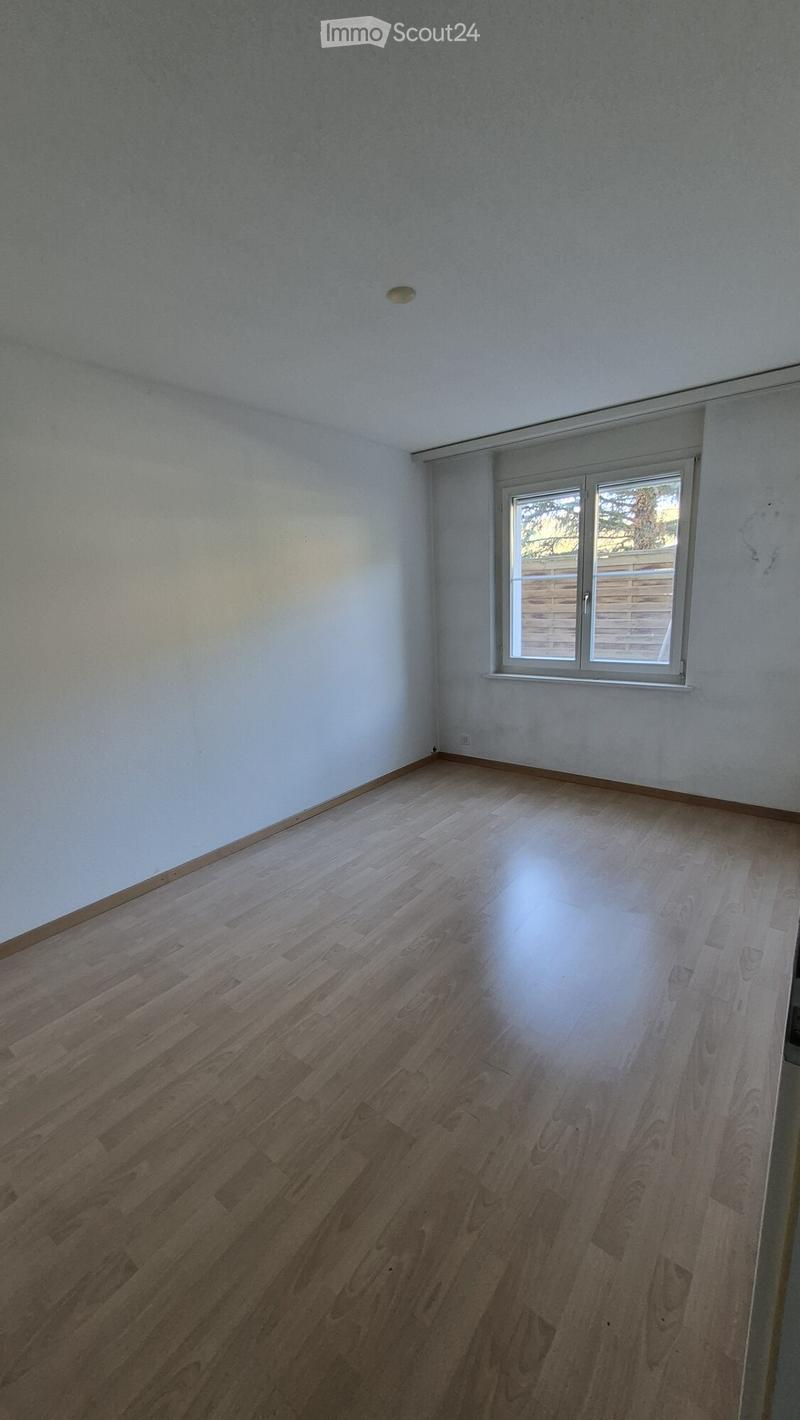 Empty room with light wooden flooring and white walls, featuring a single window with a view of a wooden fence and greenery outside.