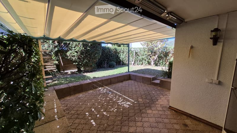 A covered outdoor patio with a brick floor, beige awning, and surrounding greenery, leading to a garden area.