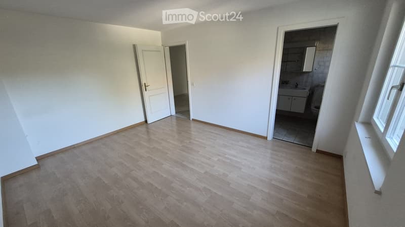 Empty room with light wooden flooring and white walls, featuring an open doorway leading to a small bathroom with a sink and toilet.