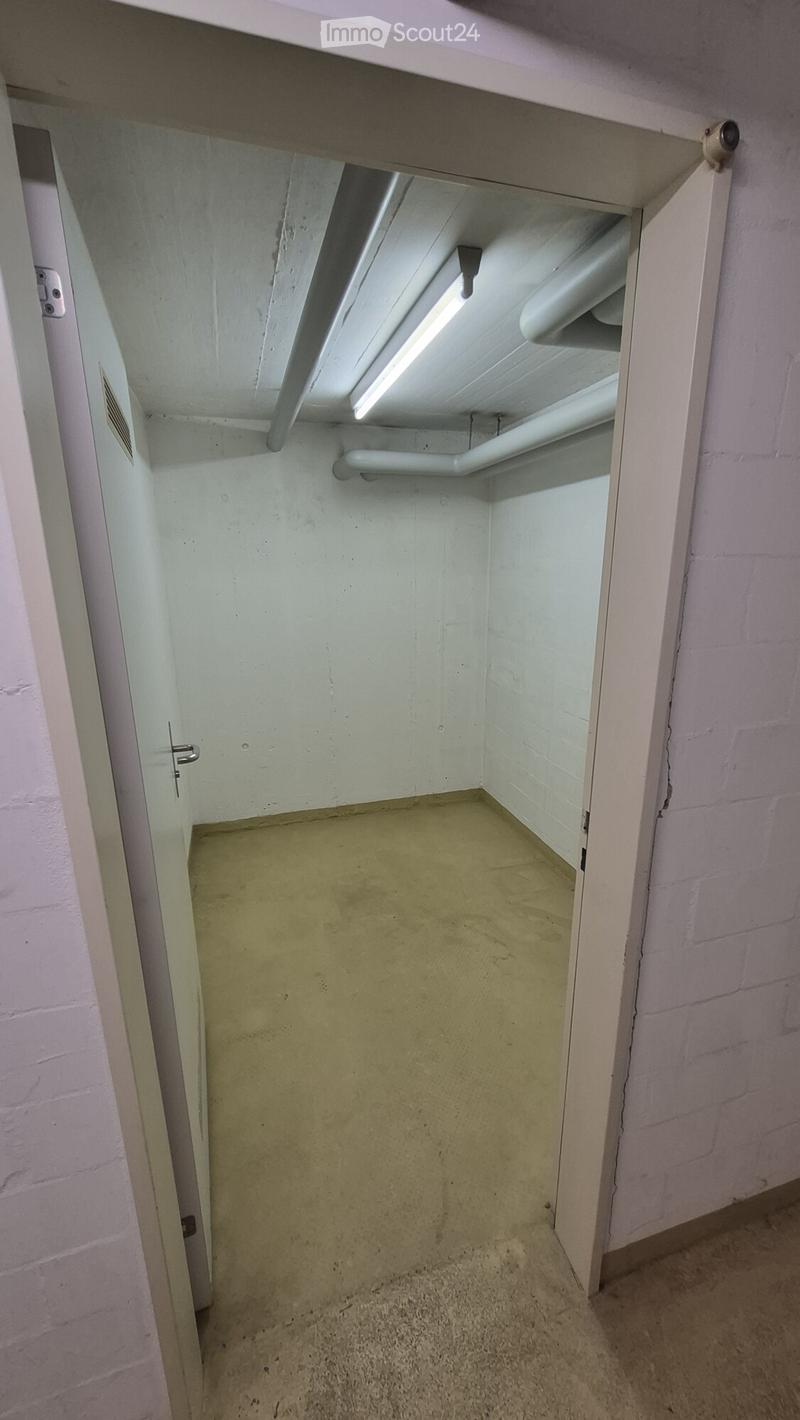 Empty room with a concrete floor, white walls, and a small window with a light fixture above it. The room has a door with a silver handle and a small vent on the wall.