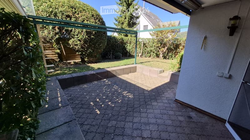 Balcony with a paved floor, low concrete wall, and a view of a well-maintained garden with trees and a wooden staircase.