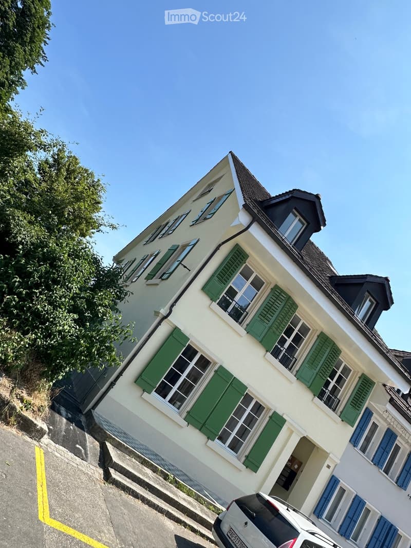 Two-story house with white walls, green shutters, and a dark roof. The house has multiple windows and a visible entrance. A car is parked in front of the house.