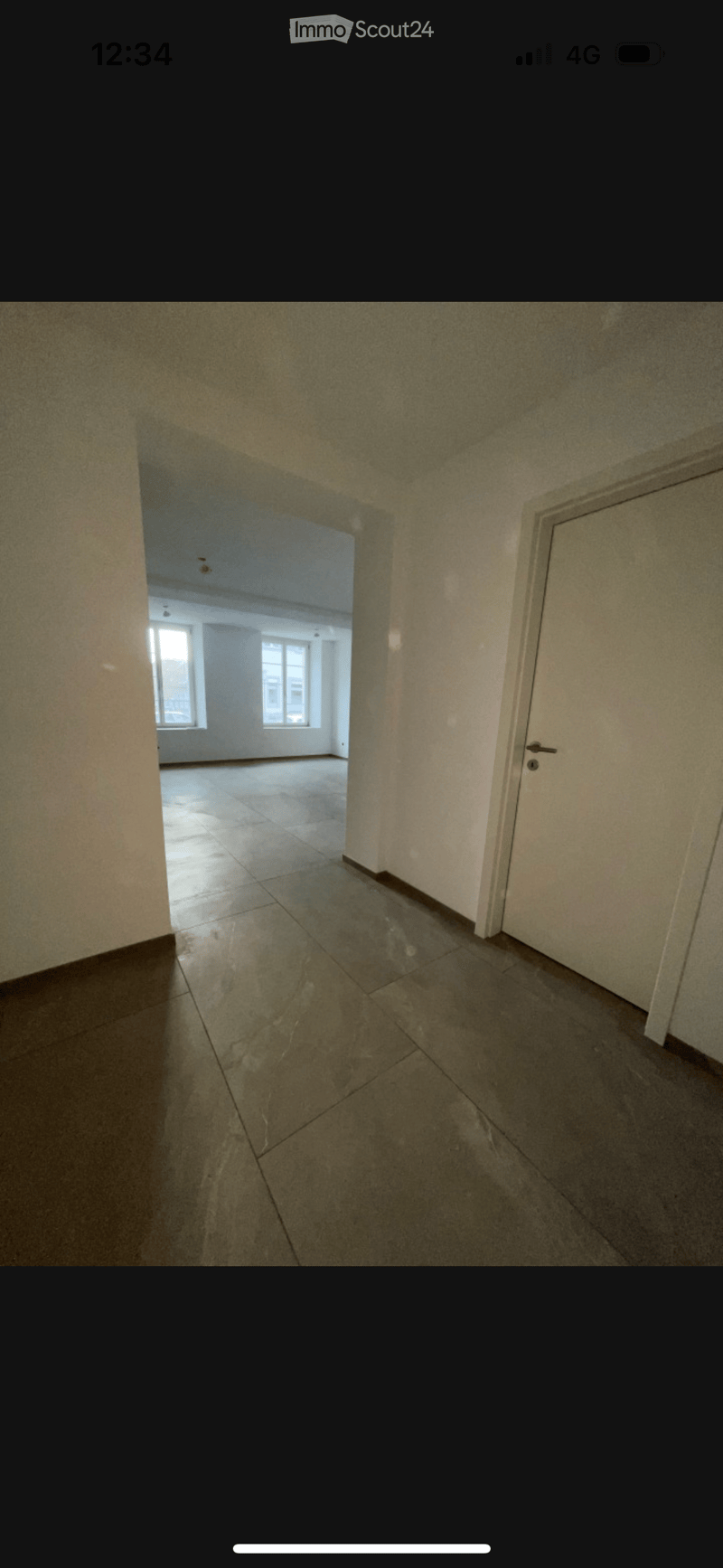 Empty hallway with tiled floor and white walls, leading to another room with windows and a tiled floor.