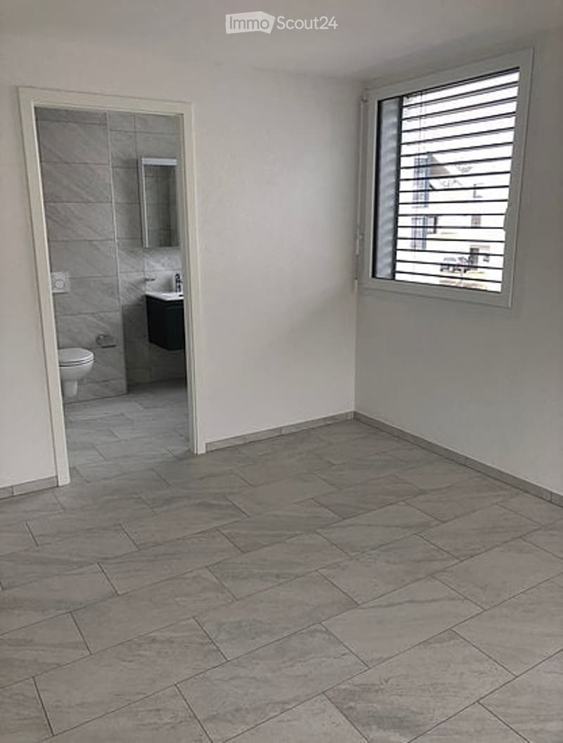 Empty room with grey tiled flooring, white walls, and a doorway leading to a small bathroom with a toilet, sink, and mirror.