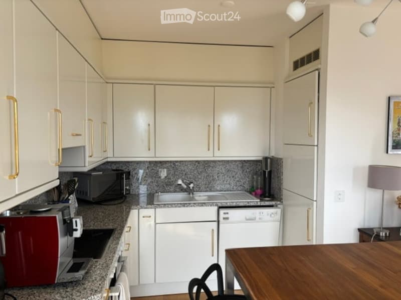 Modern kitchen with white cabinets, granite countertops, and stainless steel appliances including a microwave, toaster, and dishwasher. A small wooden dining table and chair are visible.