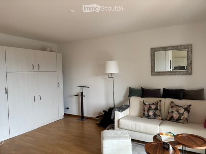 Bright living room with white walls, wooden flooring, and modern furniture including a white sofa with patterned cushions, an ottoman, a coffee table, a floor lamp, and a large mirror on the wall.