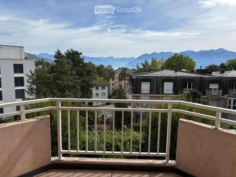 Balcony with white metal railing, overlooking a cityscape with trees and distant mountains.