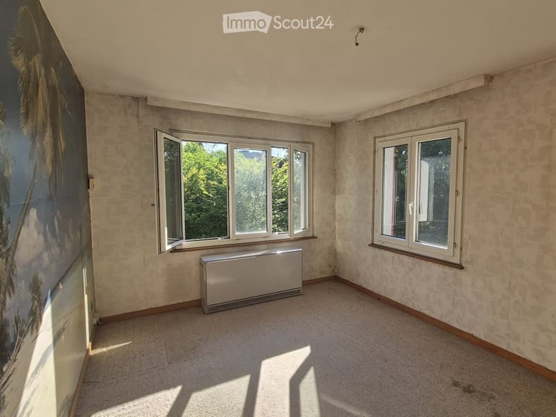 Empty room with beige walls, carpeted floor, large windows, radiator, and a mural on one wall.