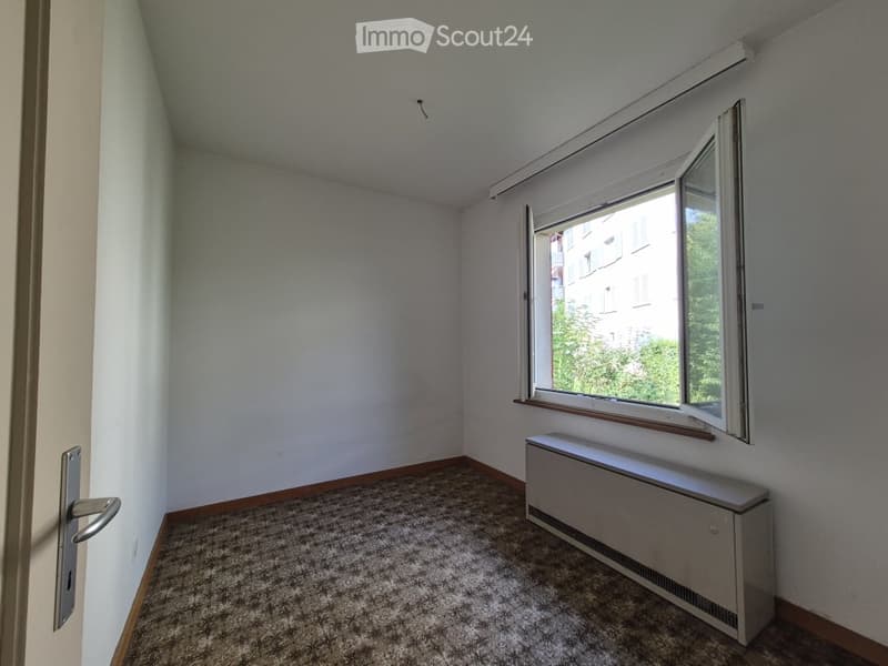 Empty room with light-colored walls, carpeted floor, a window with blinds, and a radiator below the window. No furniture or personal items are present.