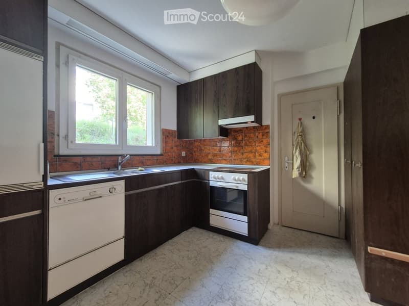Modern kitchen with dark wooden cabinets, white appliances, and a tiled backsplash. Features a sink, dishwasher, and oven, with a window providing natural light.