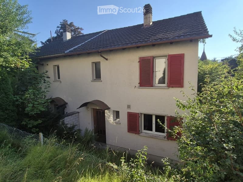 Two-story house with beige walls, red shutters, and a tiled roof. The house features multiple windows, a front door, and is surrounded by greenery.