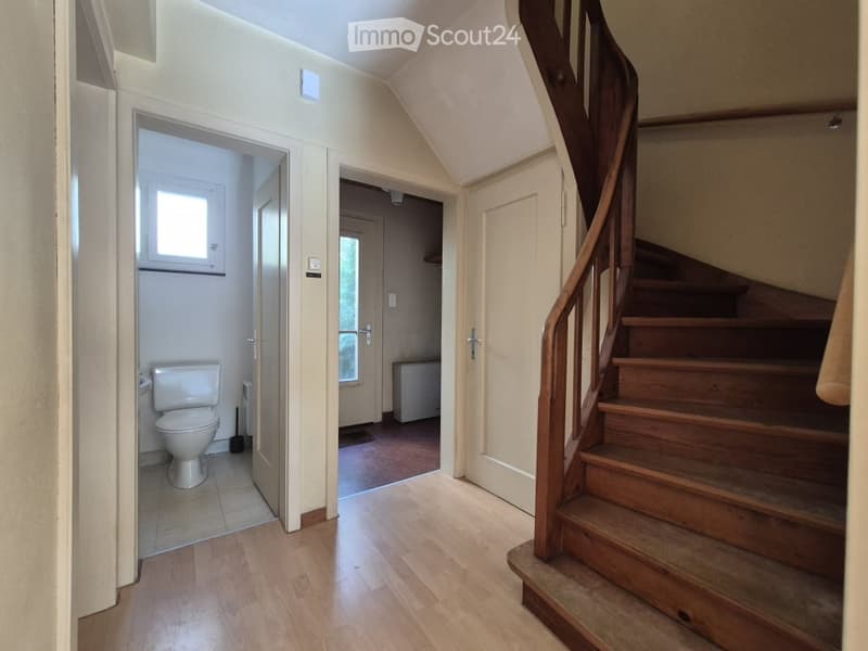 Hallway with wooden flooring, a staircase with wooden railings, and an open doorway leading to a bathroom with a toilet and window.