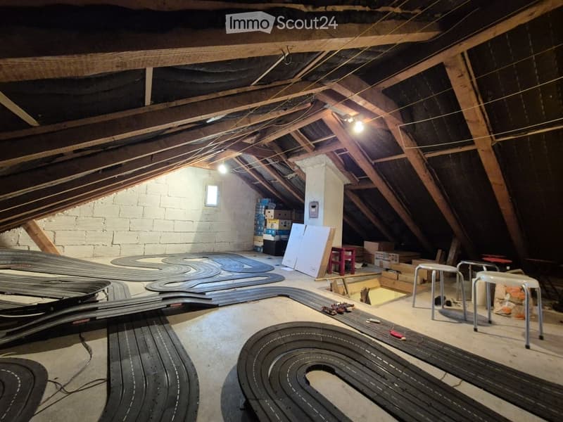 An attic space with exposed wooden beams and insulation, featuring a concrete floor and white brick walls. The room contains several RC car tracks laid out on the floor, along with storage boxes, a small table, and a few stools.