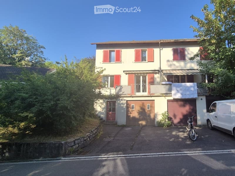 Two-story house with beige walls, red shutters, and a garage. Features a balcony on the upper floor and a driveway with a parked van and bicycle.