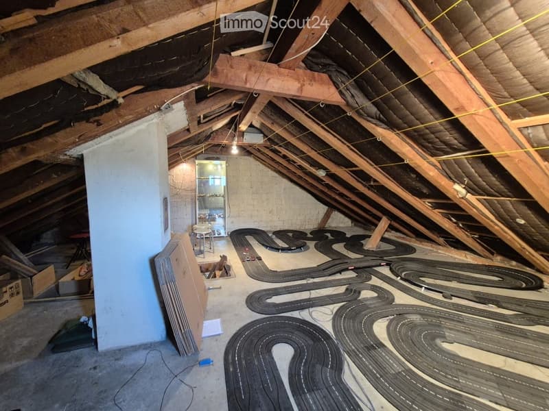 An attic space with exposed wooden beams and insulation, featuring a toy race track layout on the floor. The room includes a small doorway leading to another area, some storage boxes, and a few scattered items.