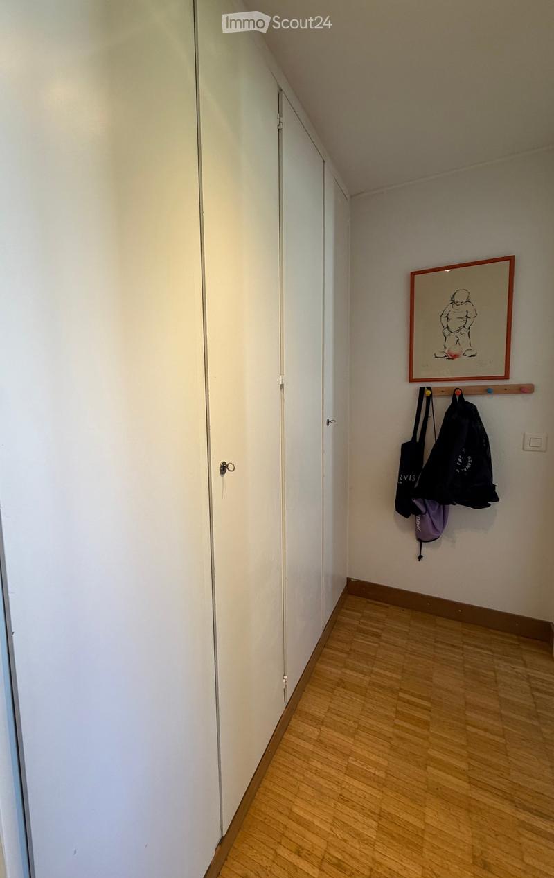 A narrow hallway with wooden flooring, white walls, and a closed white door on the left. A coat rack with two bags hangs on the right wall, and a framed picture is displayed above it.