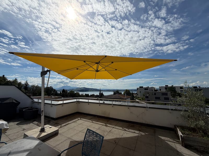 A spacious rooftop terrace with a large yellow umbrella, tiled flooring, metal railing, and a scenic view of the lake and mountains.