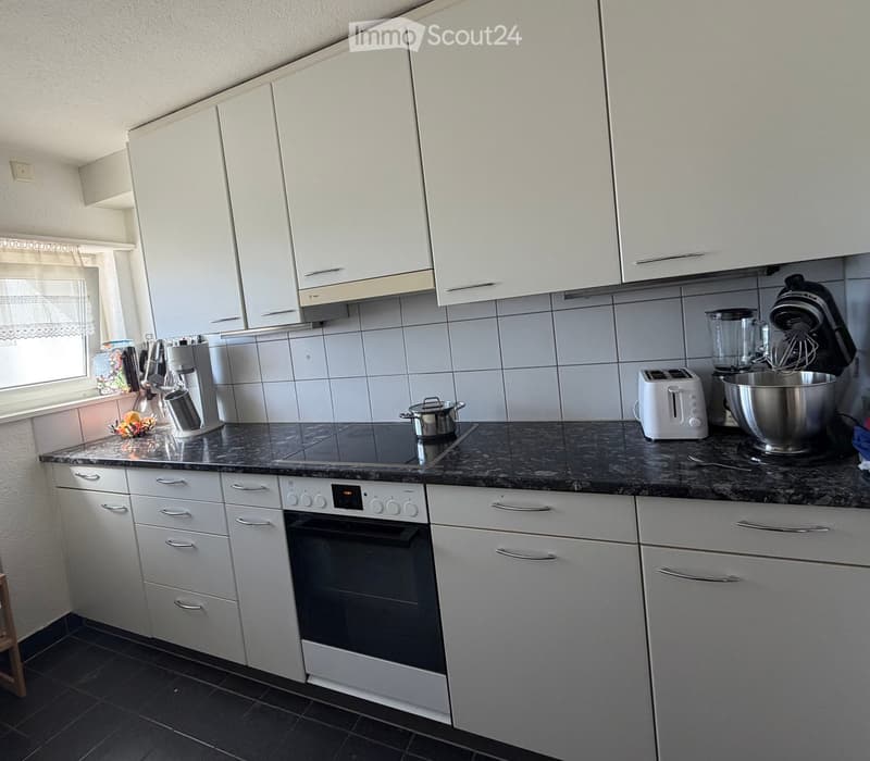Modern kitchen with white cabinets, granite countertop, and stainless steel appliances including an oven, toaster, stand mixer, coffee maker, and blender. A fruit bowl and pot are also visible.