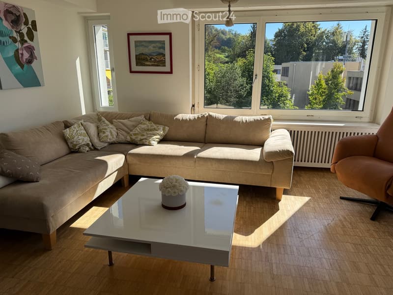 Modern living room with a sectional sofa, glass coffee table, armchair, and a flower arrangement on the table. Large windows offer a view of greenery outside.
