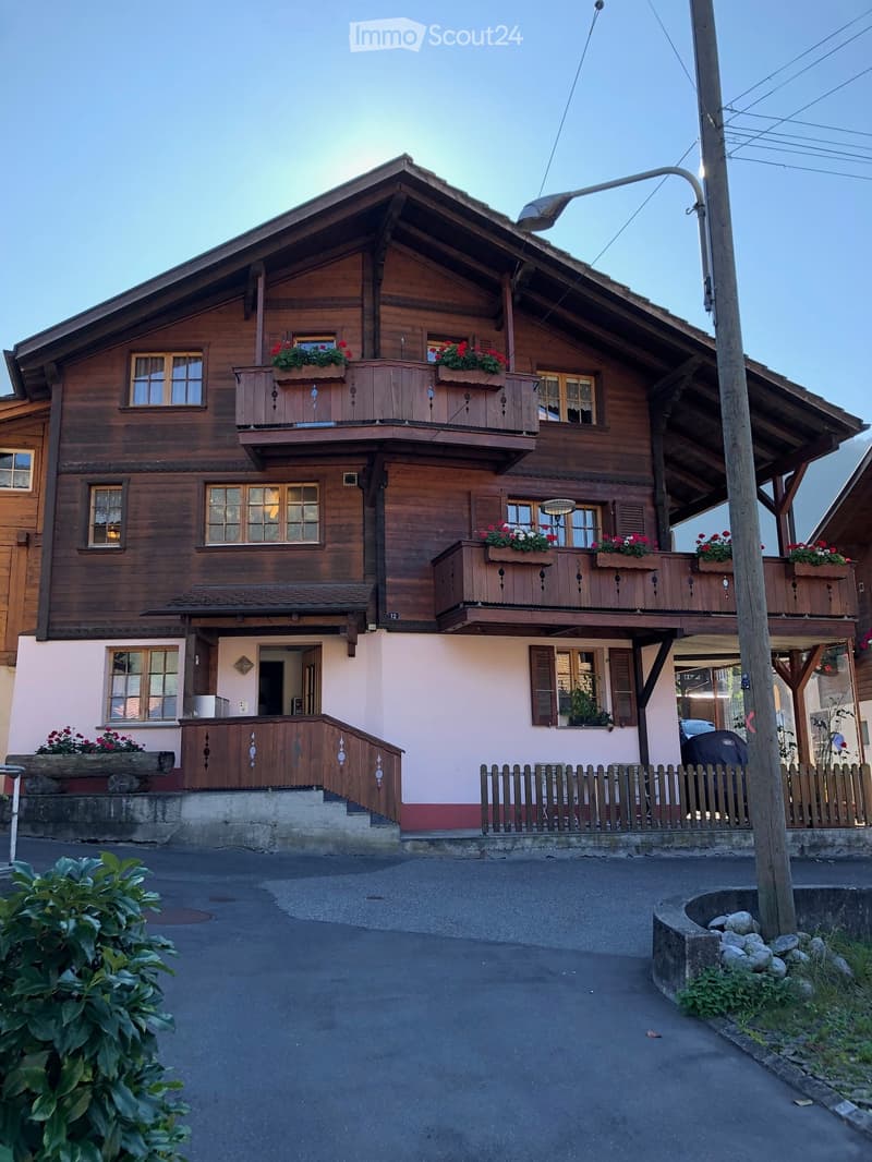 Two-story wooden house with balconies, flower boxes, and a small garden area.