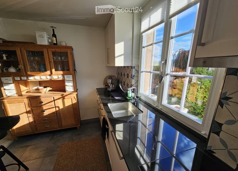 Bright kitchen with wooden cabinets, a sink, coffee maker, and various kitchen items on the counter. Large windows provide natural light and a view of greenery outside.