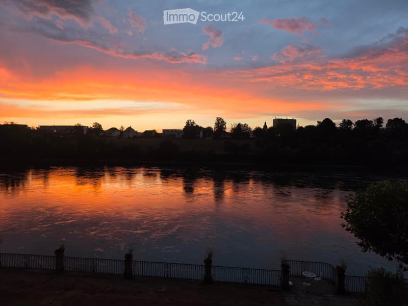 A serene riverside scene at sunset, with a calm river reflecting the vibrant orange and pink hues of the sky. A metal fence lines the riverbank, and a small seating area with a table and chairs is visible on the right.