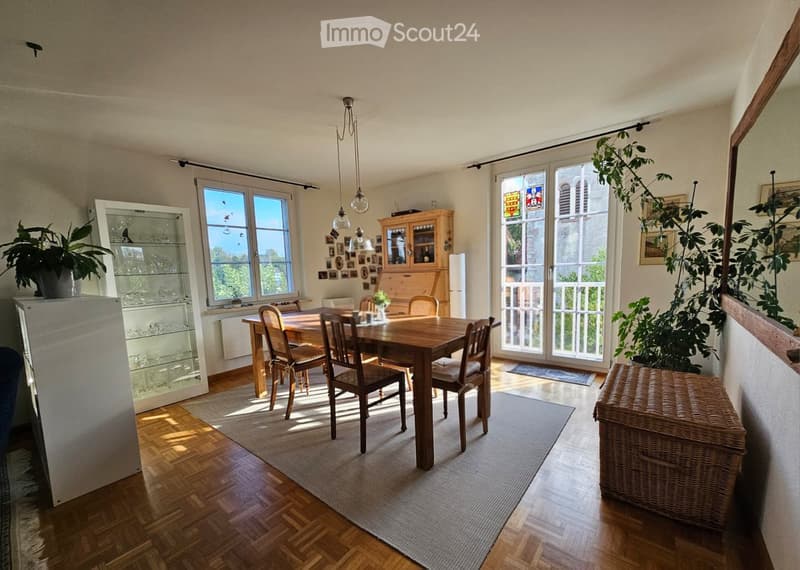 Bright dining area with wooden dining table and chairs, large windows, cabinet, refrigerator, and plants.
