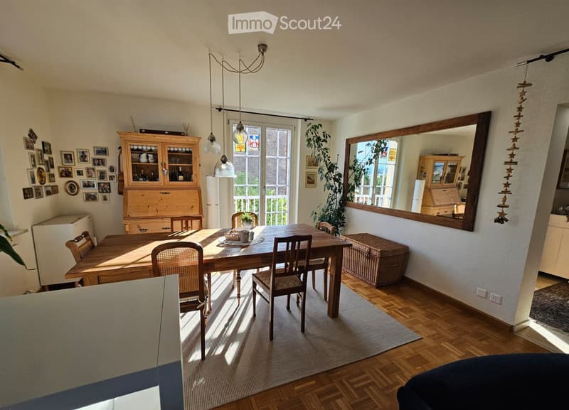 Bright dining area with wooden dining table, chairs, cabinet, large mirror, and natural light from windows.