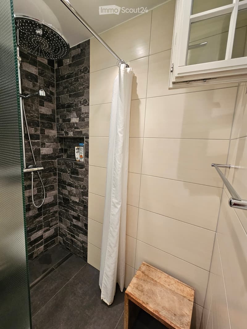 Modern bathroom with a glass-enclosed shower, dark stone tiles, a white shower curtain, a small wooden stool, and a shelf holding toiletries.