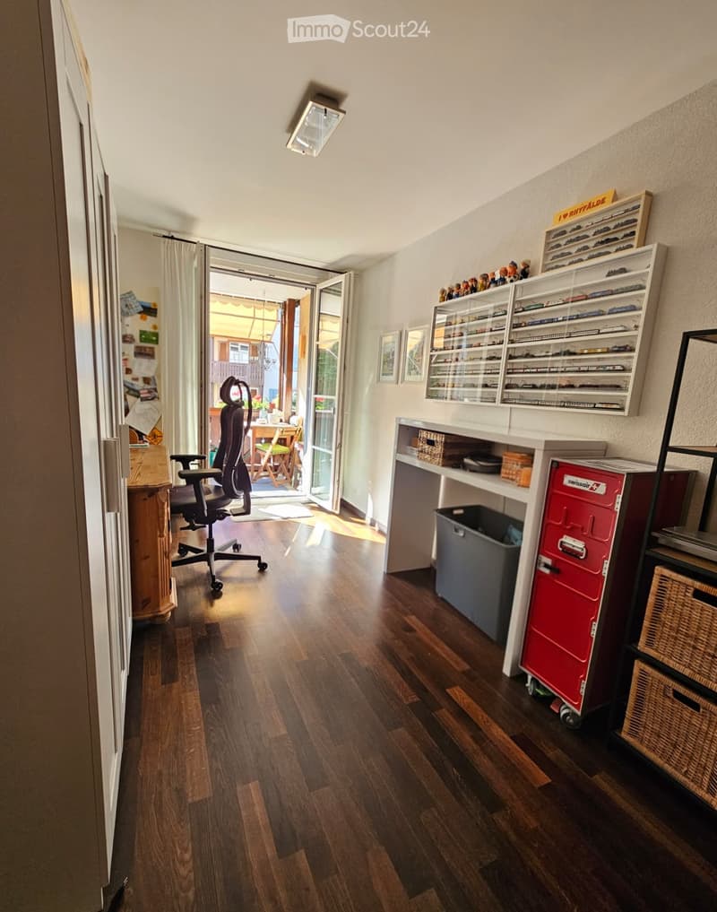 A well-lit room with wooden flooring, a large window with a curtain, a desk with shelves, a chair, and a small table outside the window.