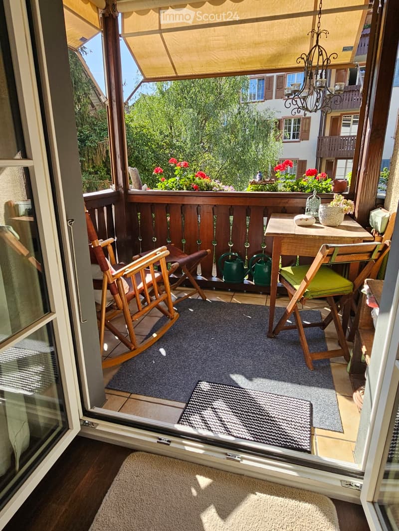 Balcony with wooden railing, small table, chairs, rocking chair, flower pots, and watering cans.