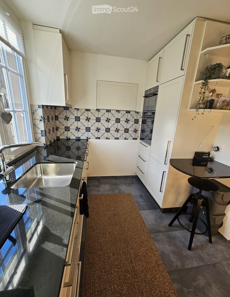 Modern kitchen with white cabinets, stainless steel sink, dark countertops, and a patterned backsplash. Includes a small stool and a rug on the floor.